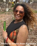 Kenya is a lighter skinned-toned woman of African descent. She has long blondish brown hair wich blows in the wind behind her, and, is wearing a black jumpsuit with yellow, green and red embellishments. She smiles brightly into the camera holding her green handled cane and gold rimmed glasses. In the background, white magnolias and green shrubbery surround her.