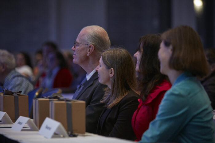 Four people sit facing away from the camera in a crowded room.