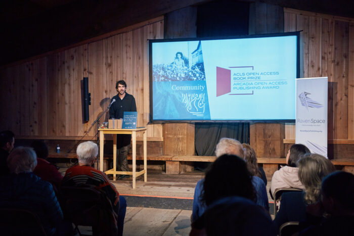 Man stands at podium beside a screen featuring the logo for the ACLS Open Access Book Prizes.