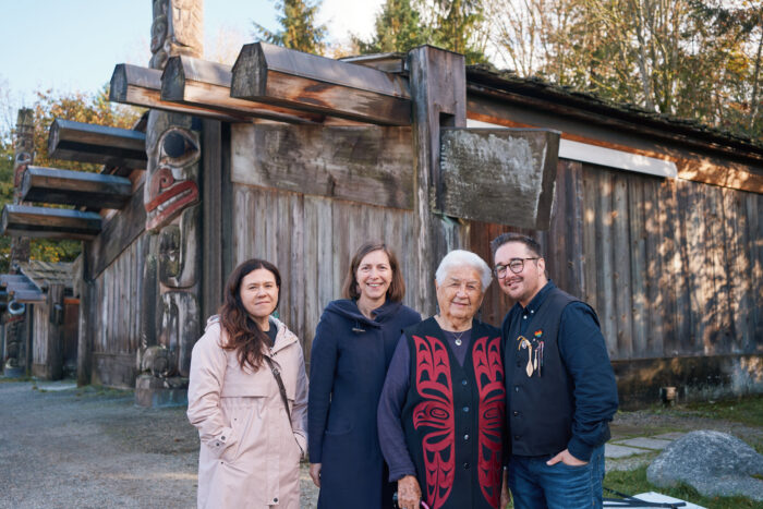 Four people stand beside a rustic structure and totem pole.