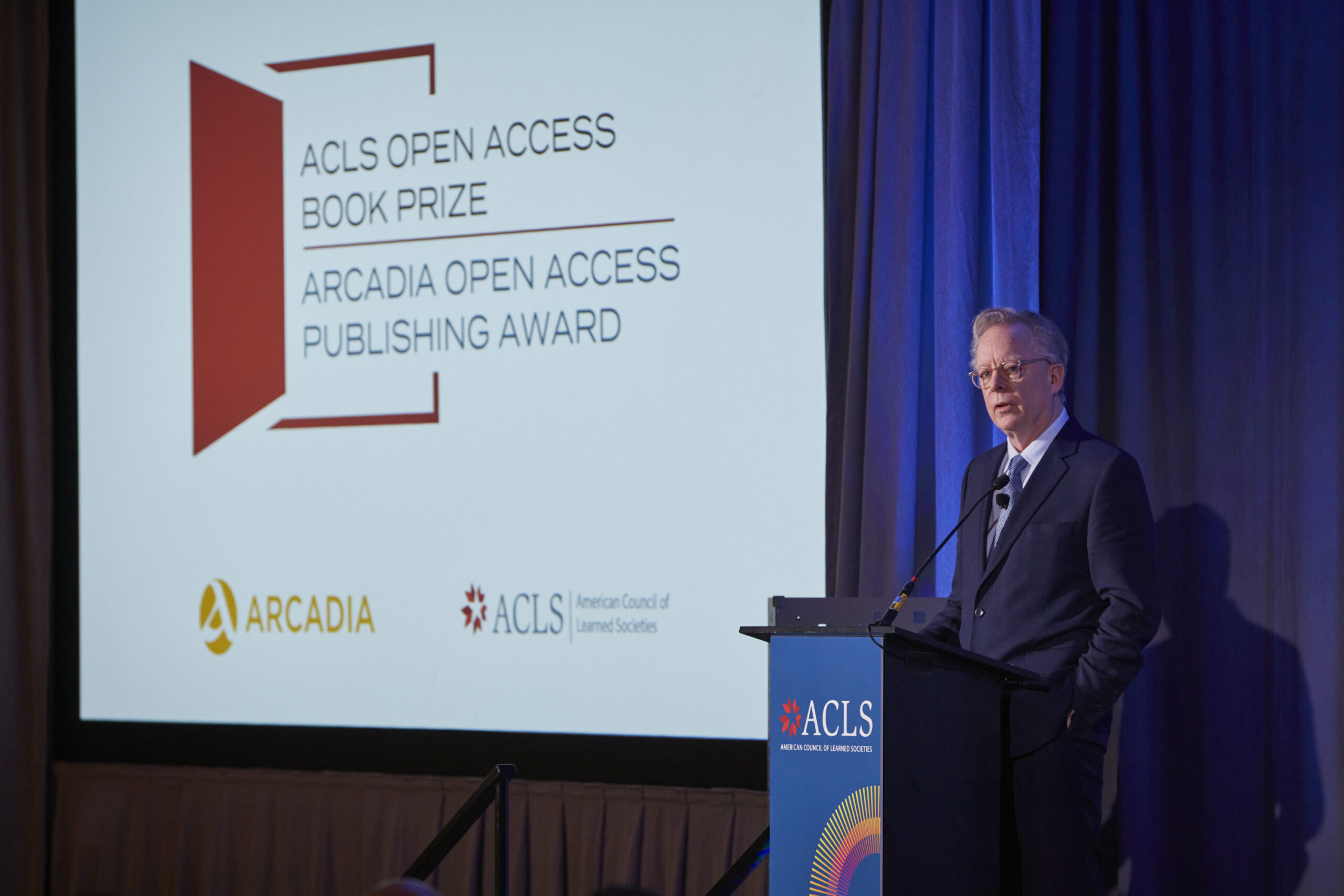 Man stands at lecturn beside screen with the logo for the ACLS Open Access Book Prize and Arcadia Open Access Publishing Award